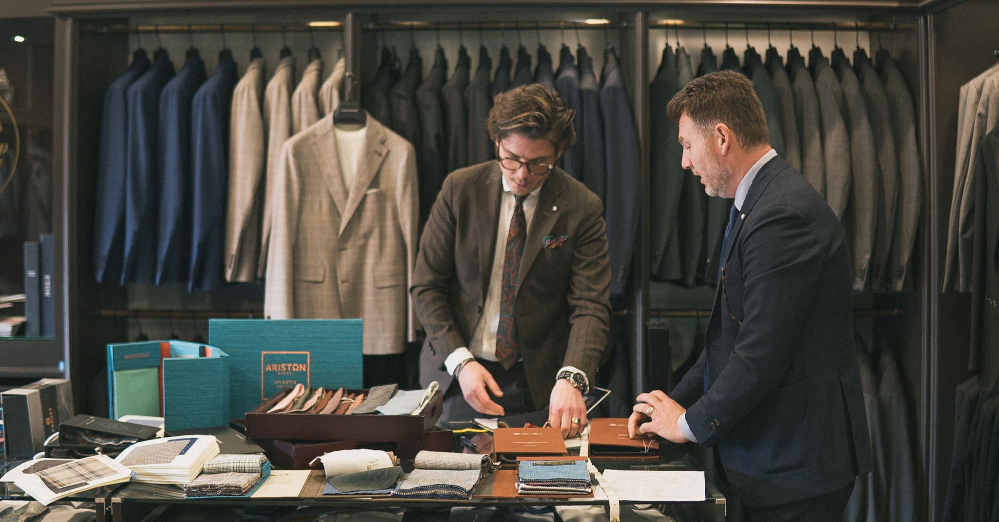 Two operators at the buying table inside Blazer for Men, fabric swatch books and supplier materials in front of a wall of jackets.